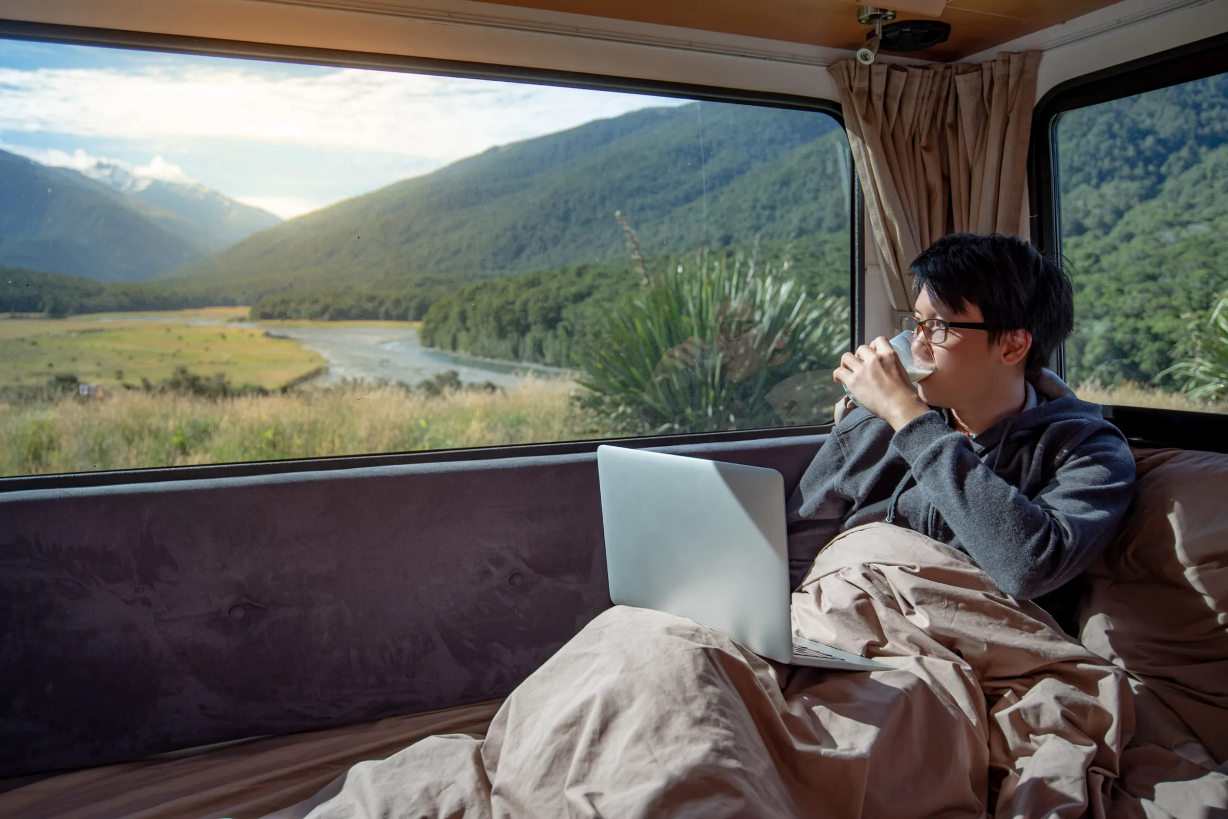 A traveler with a laptop and a hot drink, looking out over a river valley at sunrise