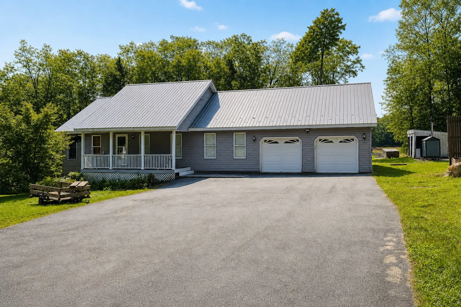 Grey ranch house with a covered porch and metal roof, framed by summer trees